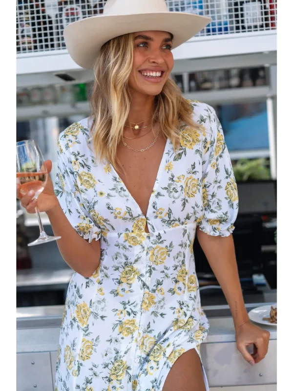 Woman in a floral dress and white hat smiling while holding a glass of wine.